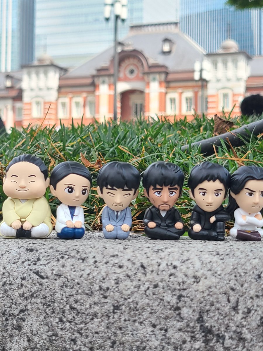 VIVANT まちぼうけの座った姿の6体のデフォルメフィギュアが石の上に横一列に並び、後ろに草と赤れんがの建物が写っている