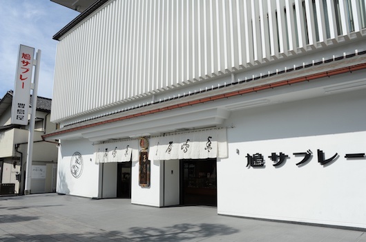 鳩サブレーVIVANT缶の白い外壁に黒い文字で鳩サブレーと書かれた豊島屋の本店の建物。入口に白いのれんが掛かる