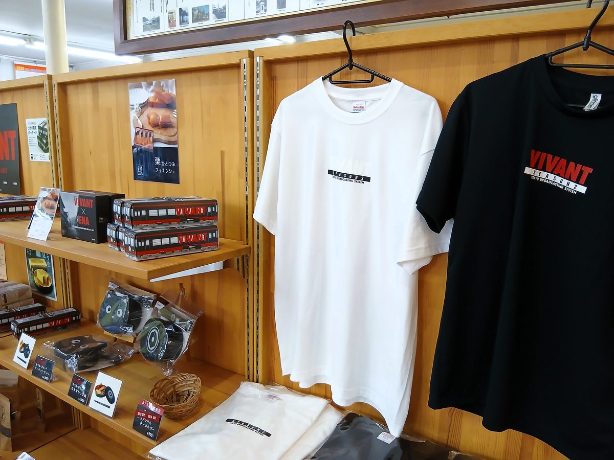 VIVANTの岐阜ロケ地の観光案内所のグッズ売り場。壁のハンガーに掛かった白と黒のTシャツと、左側の棚に並ぶ箱や袋入りの小物