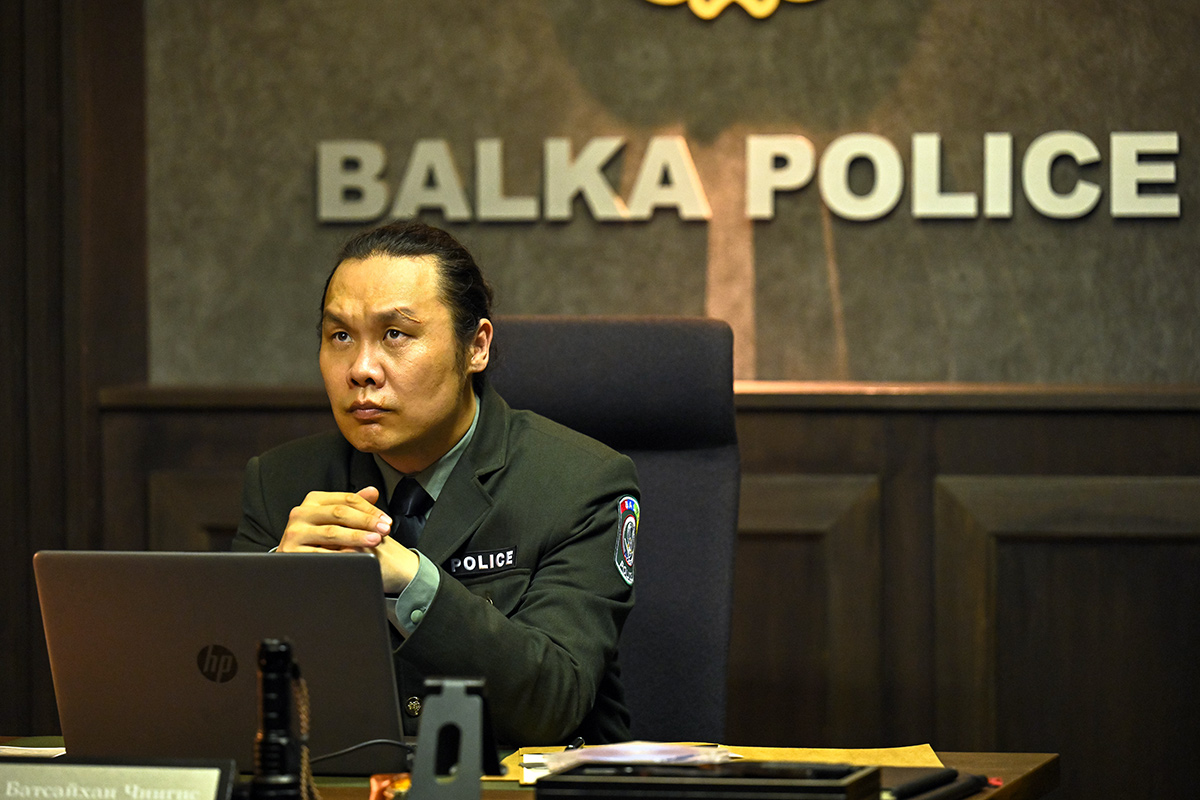 バルカ共和国の「BALKA POLICE」の文字の壁の前で机に向かう制服姿の人物（第15話の場面写真）