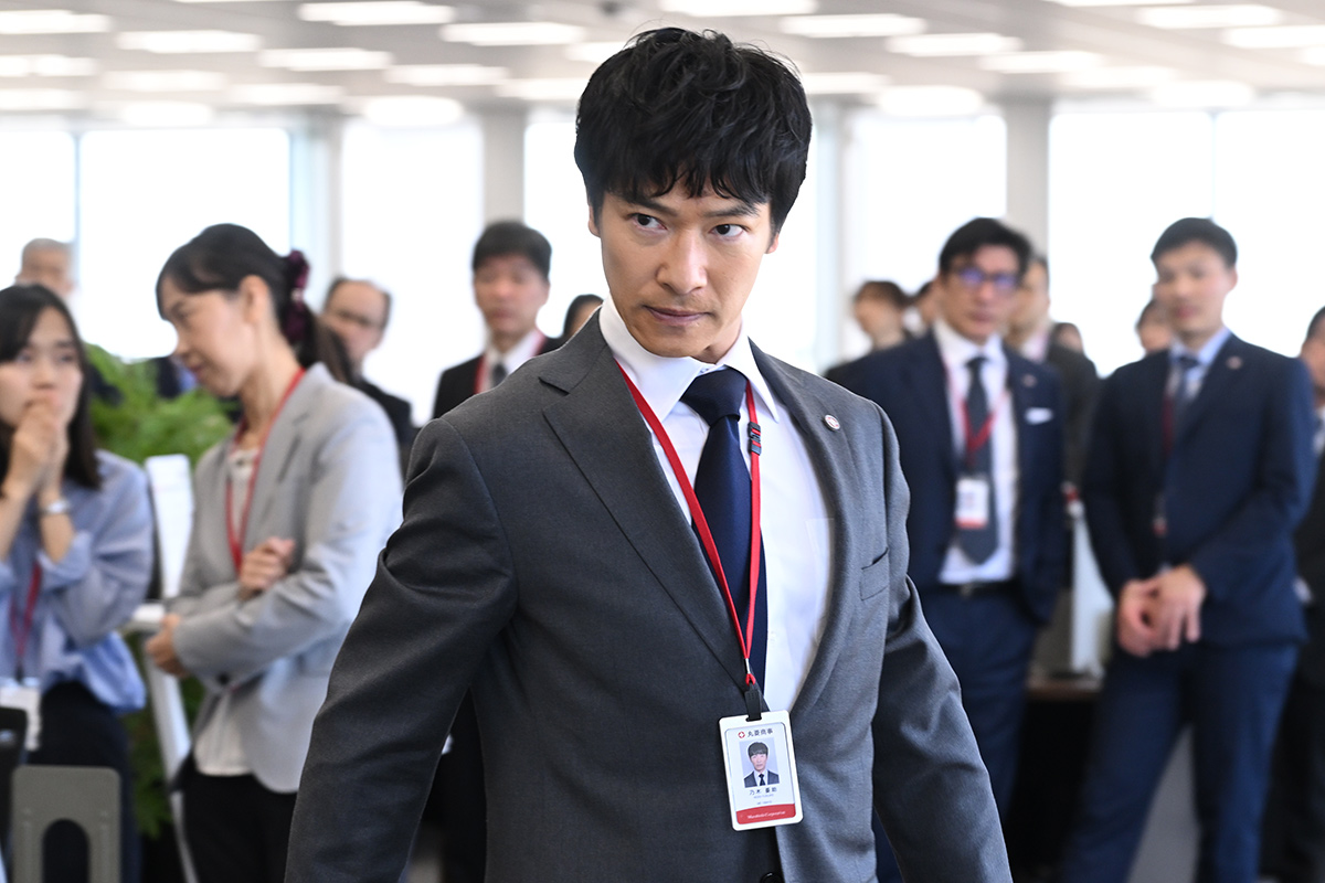 別班と公安のオフィスで赤いひもの社員証を下げて歩くスーツ姿の男性（社員証に「丸菱商事」の文字）