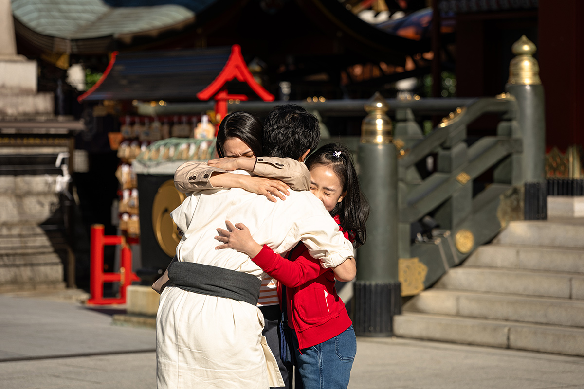 VIVANTの神社の境内で、白い服の人物がベージュの上着の女性と赤い服の女の子をまとめて抱きしめている場面