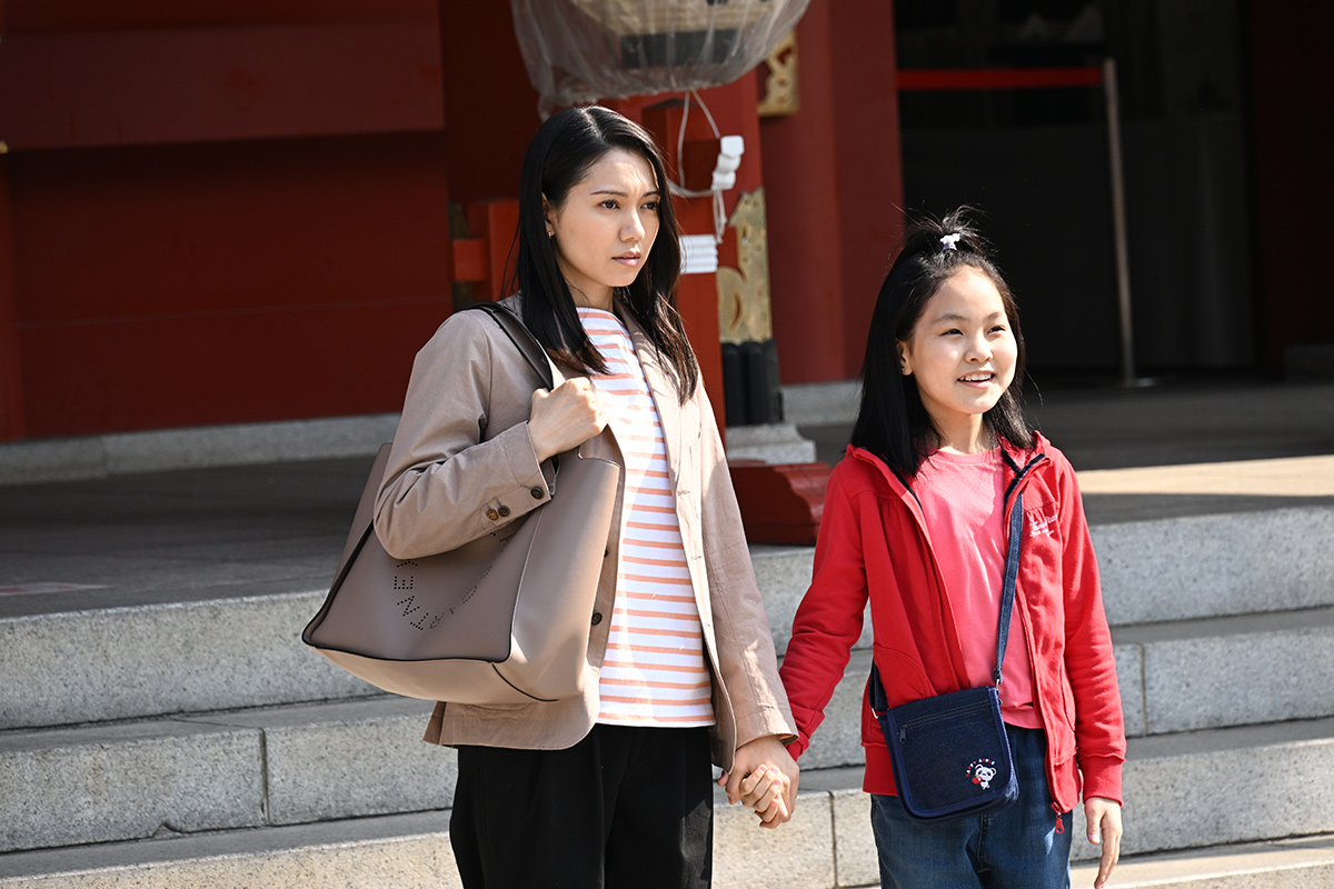 VIVANTの朱色の建物の前の石段で、ベージュの上着の女性と赤い上着の女の子が手をつないでいる場面
