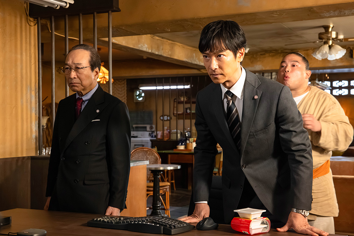 VIVANT第13話の眼鏡を掛けたスーツ姿の男性と、机に両手をついたスーツ姿の男性が並び、その後ろにベージュの服の男性が立っている場面