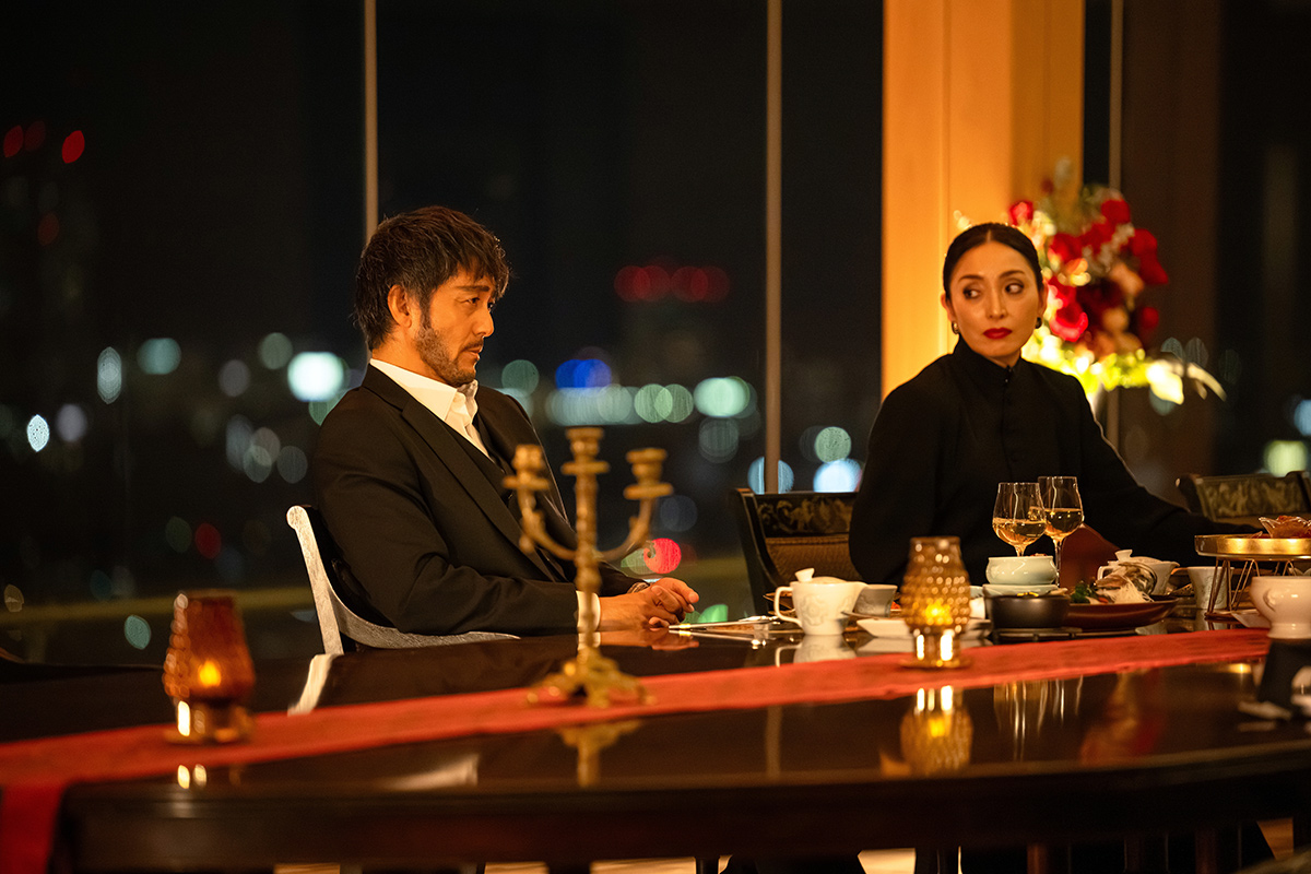 VIVANT第14話の夜景の見える窓辺で、黒いスーツの男性と黒い服の女性が、燭台の置かれたテーブルに着いている場面