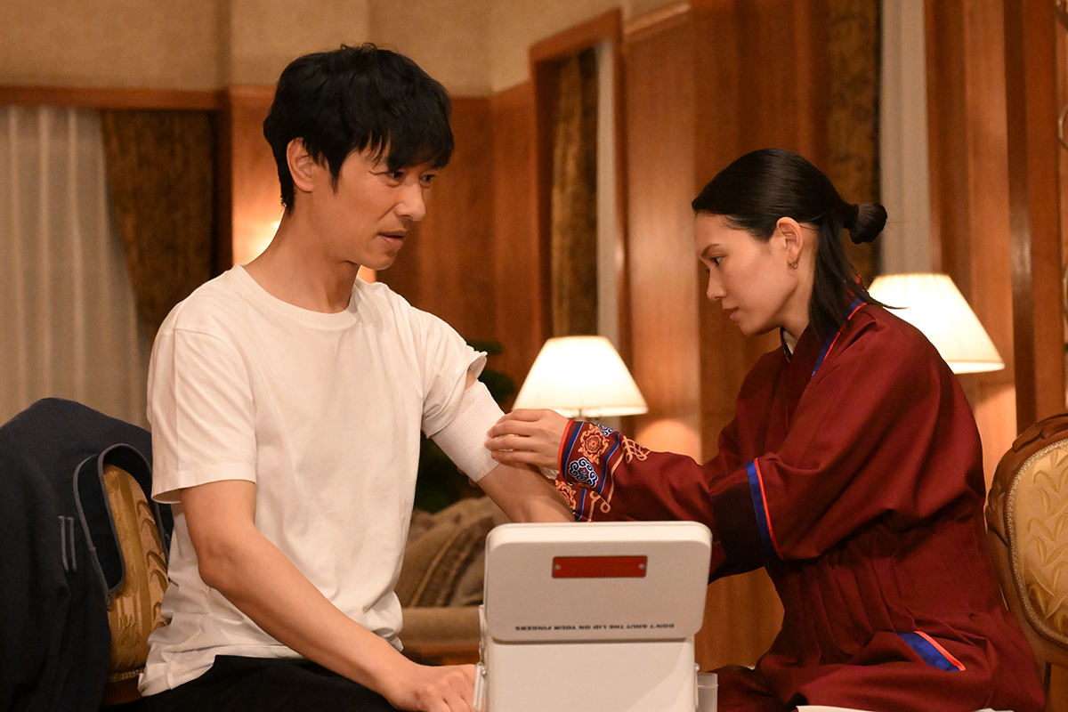 VIVANT第2話の赤い服の女性が、白いTシャツの男性の腕に白い帯を巻いている場面