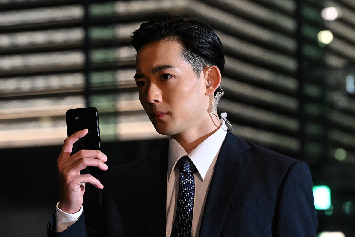 VIVANT第4話の夜の建物の前で、耳にイヤホンをつけたスーツの男性が、手に持ったスマートフォンを見ている場面