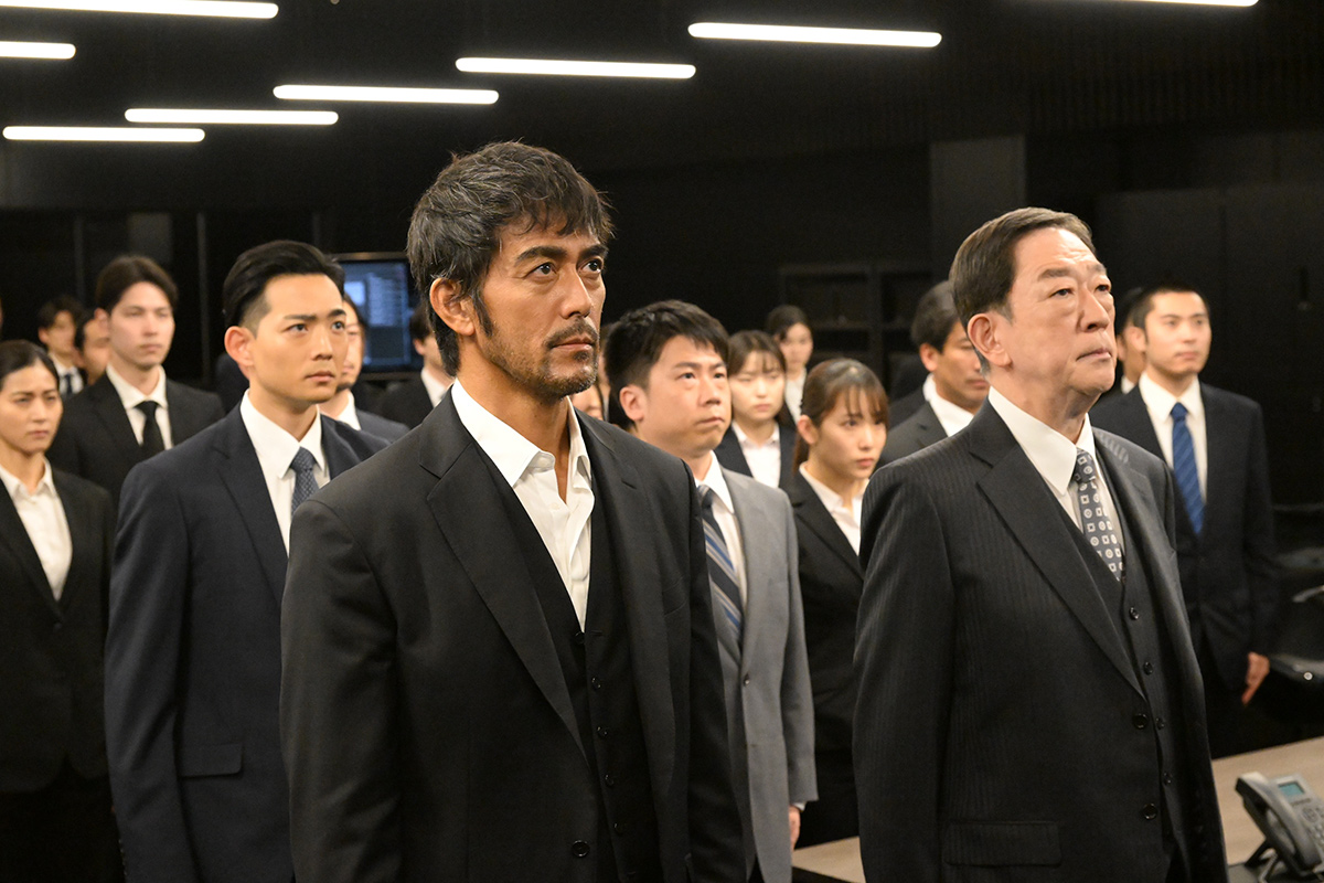 VIVANT第4話の天井に細長い照明が並ぶ暗い部屋で、スーツ姿の大勢の人が同じ方向を見上げるように立っている場面