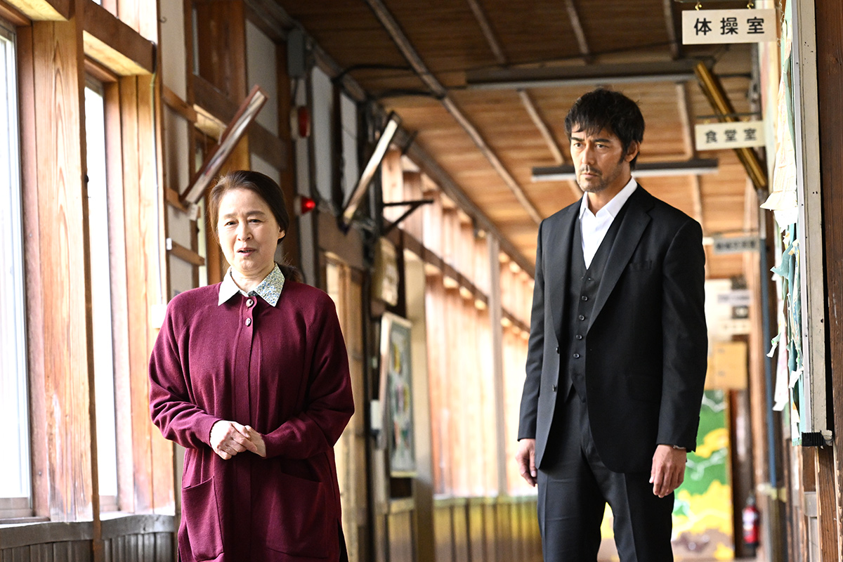 VIVANT第5話の「体操室」「食堂室」の札が下がる木造の廊下で、えんじ色のカーディガンの女性と黒いスーツの男性が立っている場面