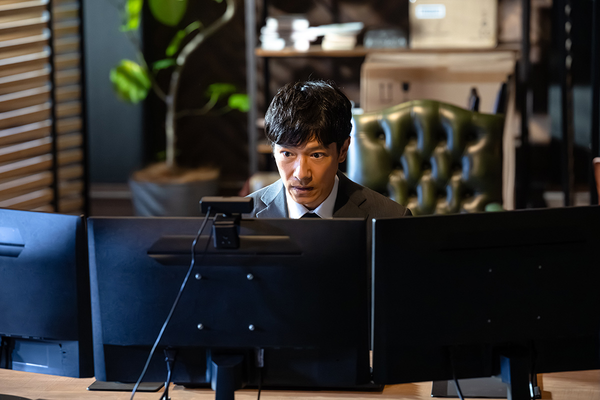 VIVANTのハヤトの横に並んだモニターの向こうで画面を見つめるスーツ姿の人物の場面写真