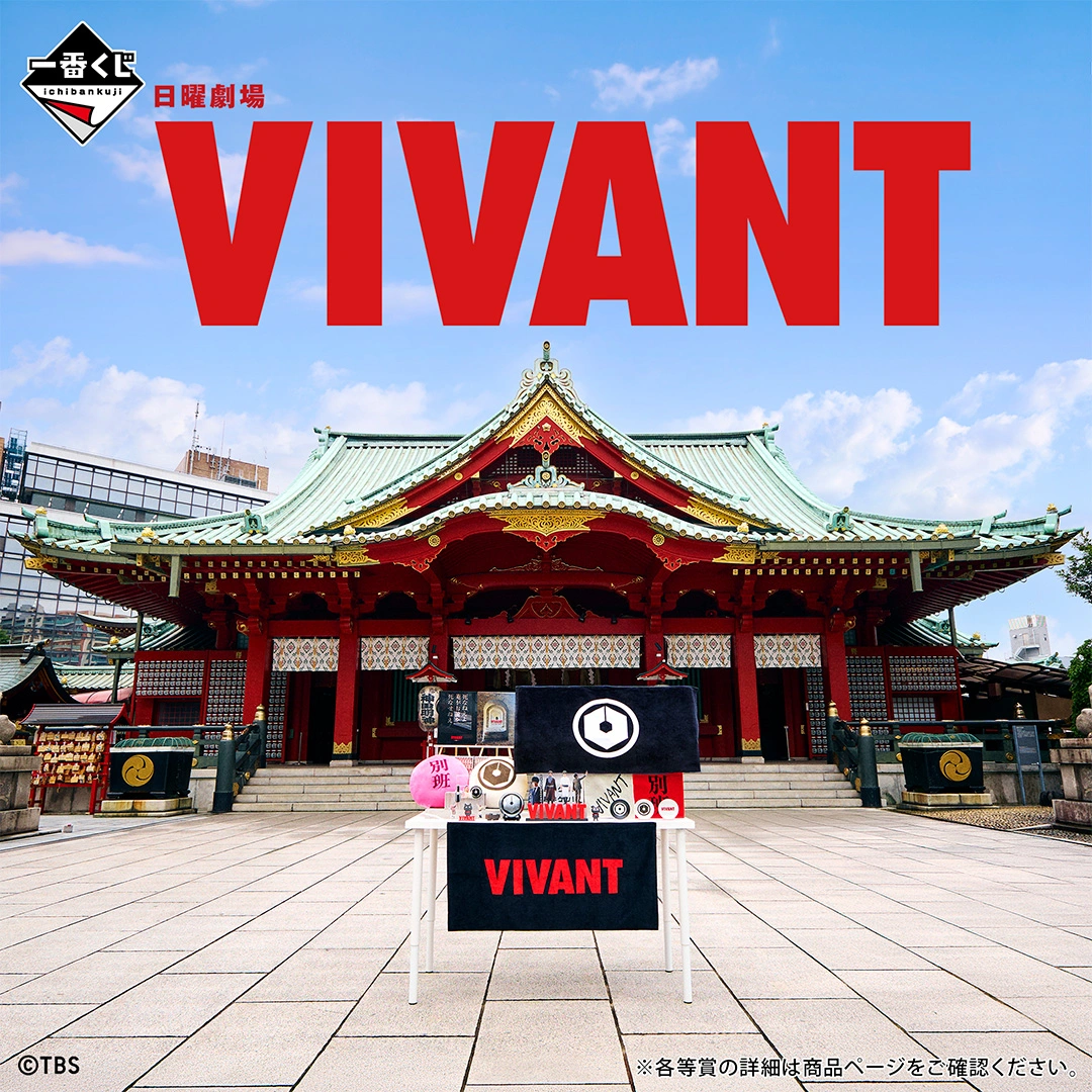 VIVANT一番くじの朱色の柱の建物の前の石畳に置かれた白い台。前面に黒い地に赤い「VIVANT」の文字のタオルが掛かり、上にピンク色の「別班」の丸いクッションなどが並ぶ