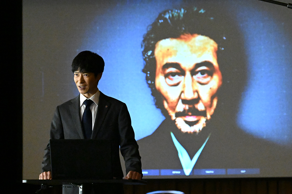 黒須のスクリーンに映った白いひげの男性の顔の前で、スーツの男性が演台に立つ場面(第7話の場面写真)