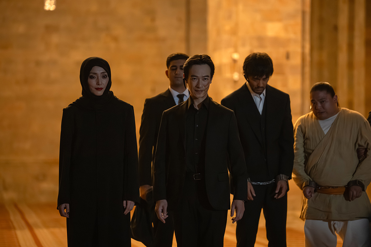 VIVANTの石の壁の建物の中に並ぶ、黒い布で頭を覆った人とスーツの男性たちとベージュの服の男性