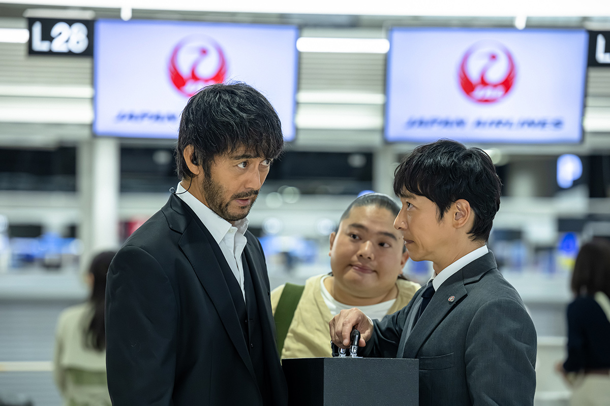 VIVANTの千葉の第11話の場面写真。「L28」の表示が見える空港のカウンターの前で向き合う3人