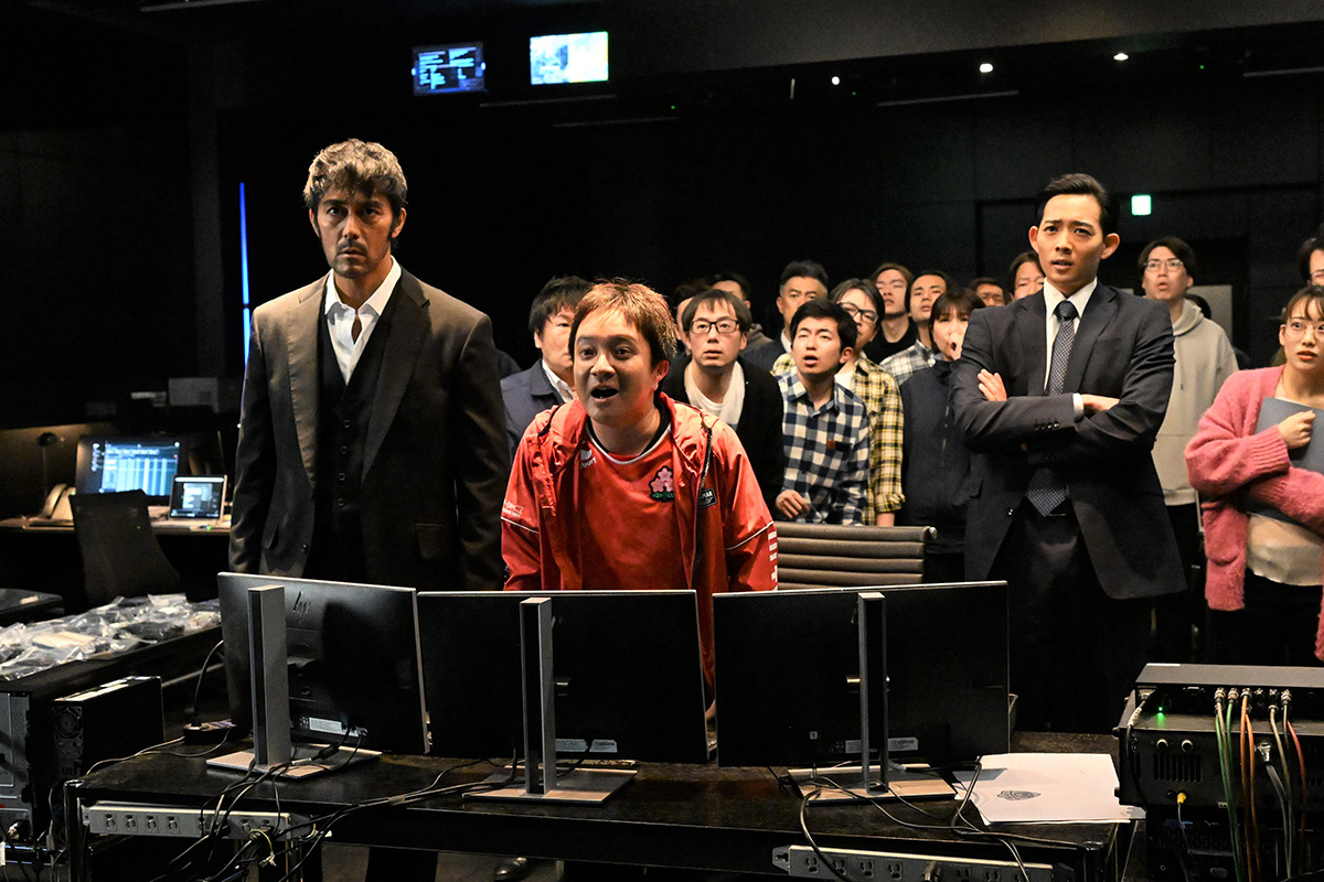 VIVANTの浜松の第4話の場面写真。暗い部屋でモニターの並ぶ机の向こうに大勢が立ち、同じ方を見る姿