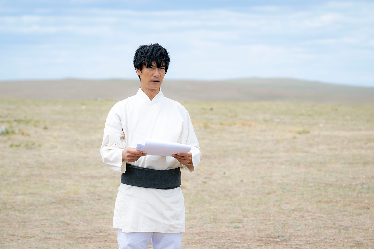 VIVANTの浜松の第9話の場面写真。平原と低い丘を背に、白い服に黒い帯の男性が白い紙を持って立つ姿