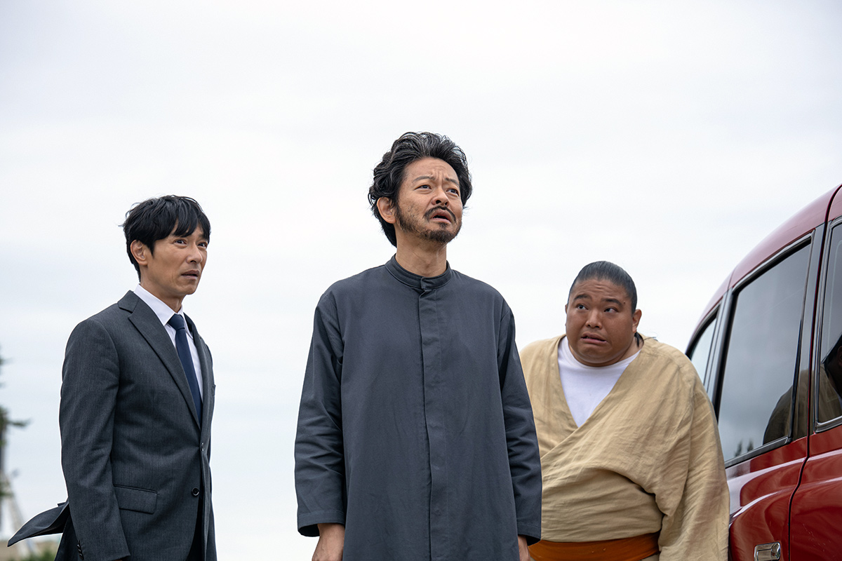 VIVANTの浜松の第12話の場面写真。曇り空の下、赤い車のそばに並んで立つ3人の男性