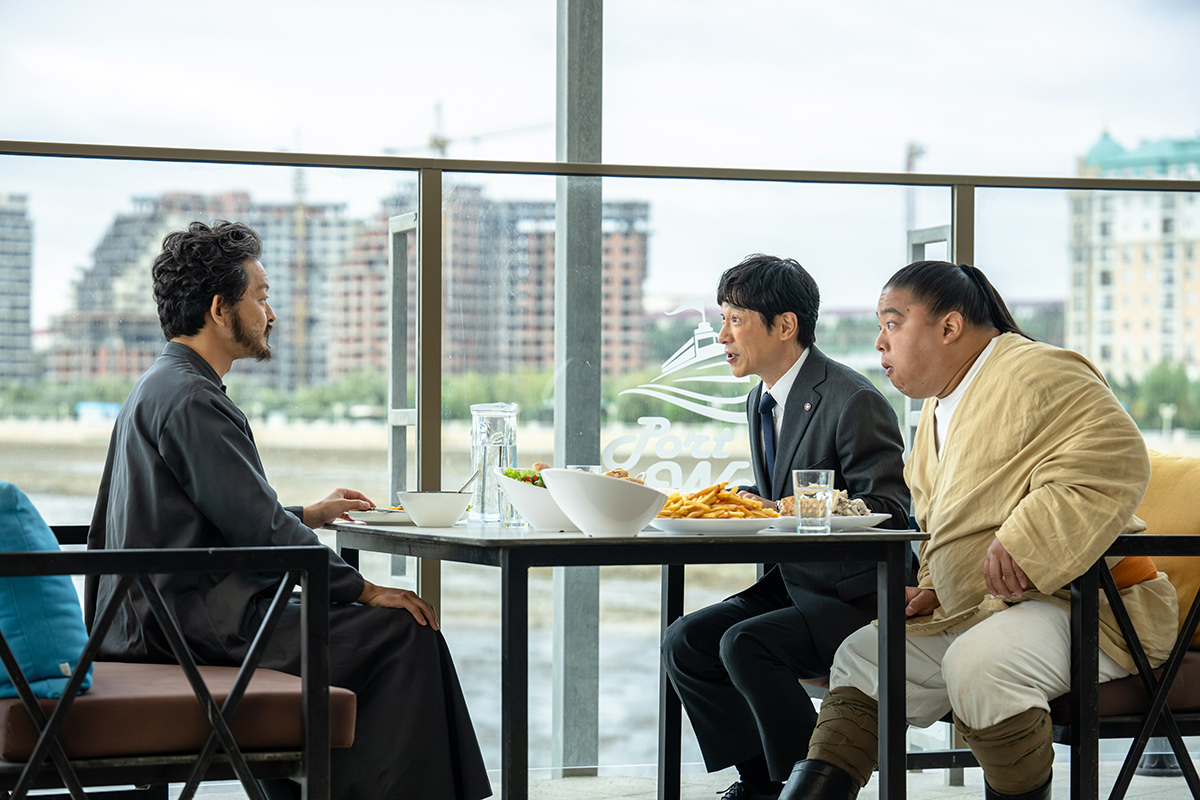 VIVANTの静岡の第12話の場面写真。高い建物が並ぶ水辺を背に、テーブルで食事を囲む3人の男性