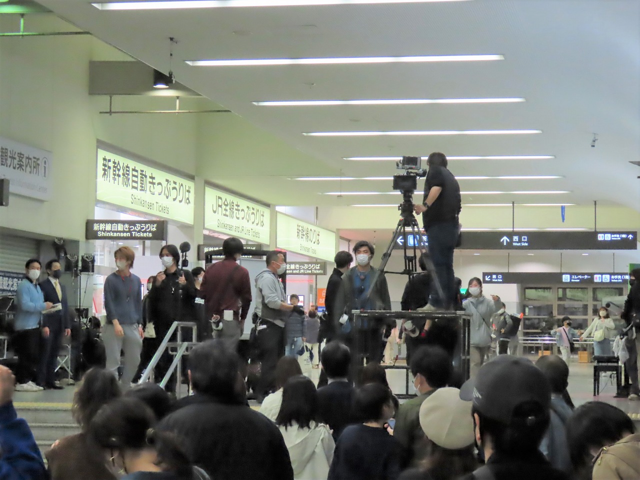 VIVANTの豊橋ロケ地のきっぷ売り場の看板が並ぶ構内で、台の上からカメラを構える人と、その前に集まった大勢の人