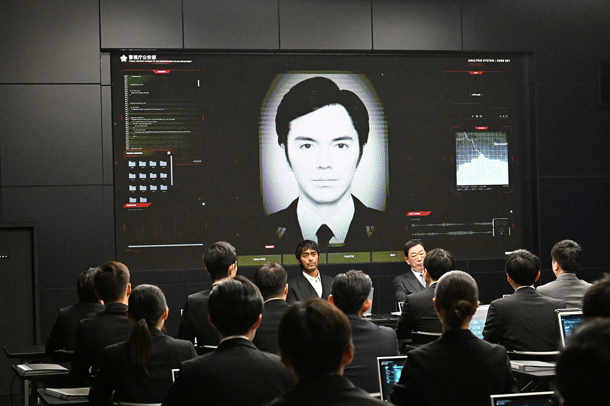 モニターの「警視庁公安部」と表示された大きな画面に男性の白黒の顔写真が映り、その前でスーツ姿の大勢の人が机に向かう場面