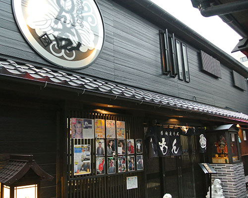 VIVANTのもんじゃの瓦屋根の軒の下に黒いのれんが下がり、入口の脇に人物の写真が何枚も貼られたもん吉の店
