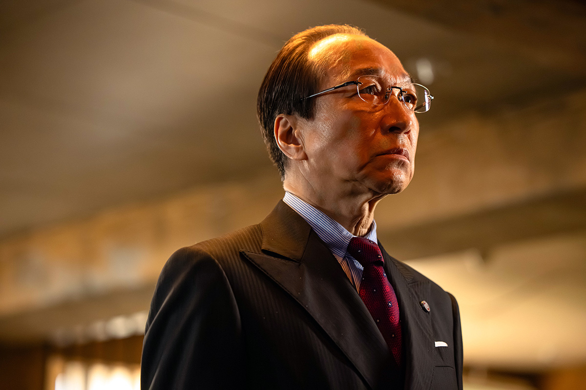 長野専務の第13話の場面写真。眼鏡をかけ、えんじ色のネクタイを締めたスーツ姿の年配の男性が遠くを見ている