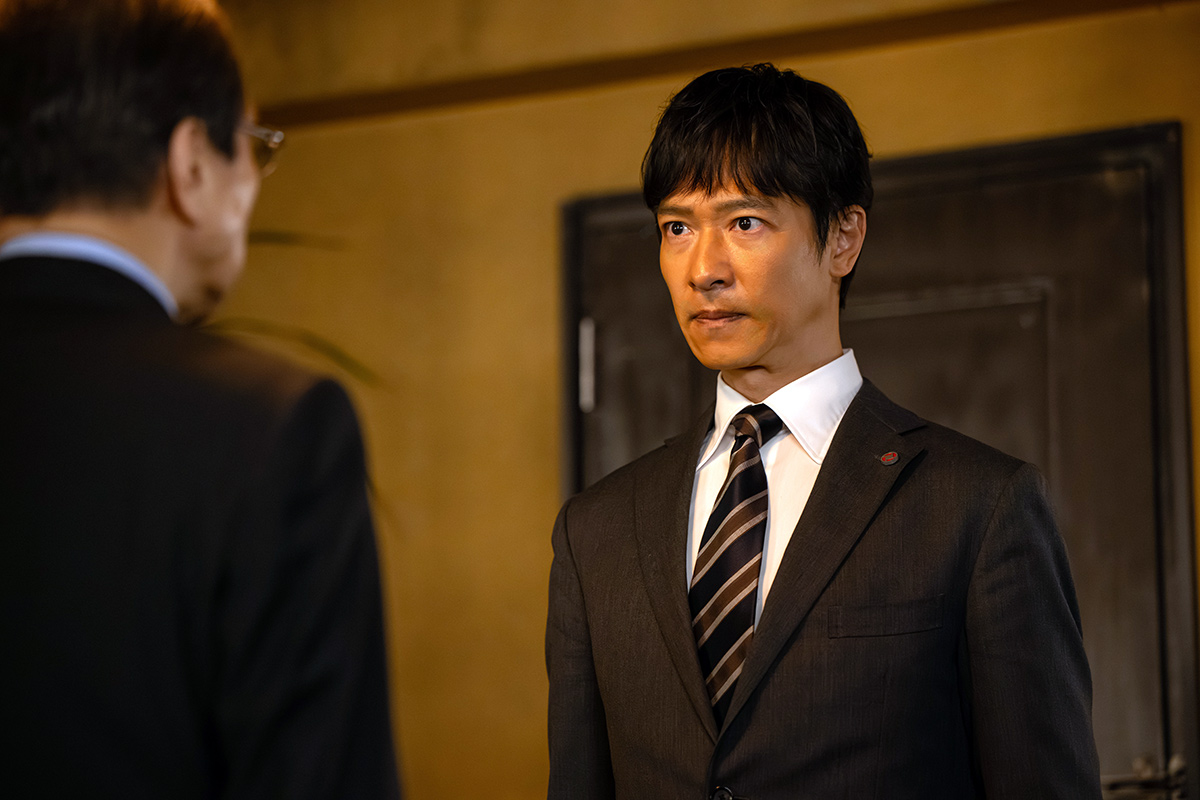 長野専務の第13話の場面写真。ストライプのネクタイのスーツ姿の男性が、手前の眼鏡の人物をまっすぐ見つめている