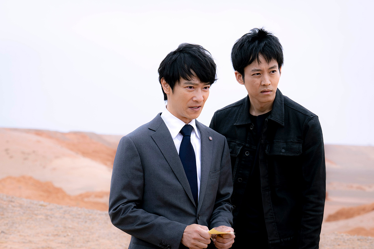 乃木の赤茶けた砂地の丘を背に並んで立つ、スーツの男性と黒いジャケットの男性(第6話の場面写真)
