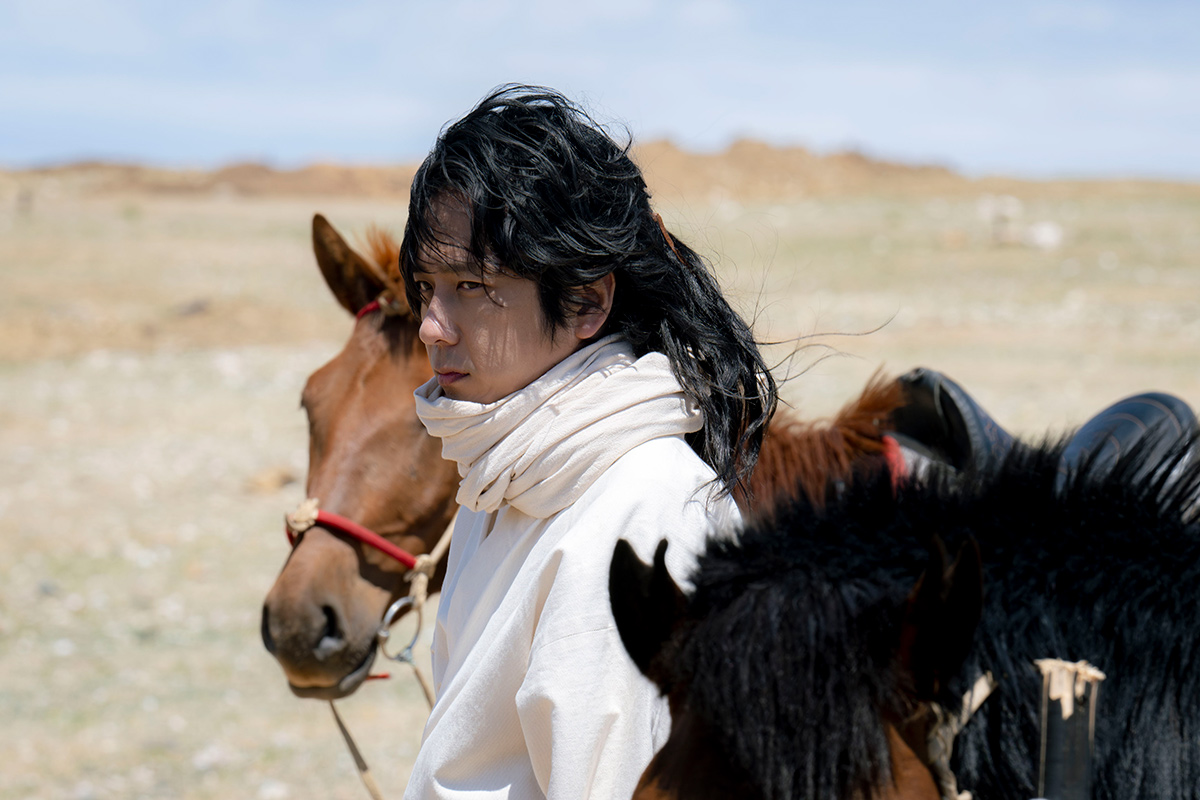 ノコルの第9話の場面写真。白い服の長い髪の男性が、草原で馬のそばに立っている