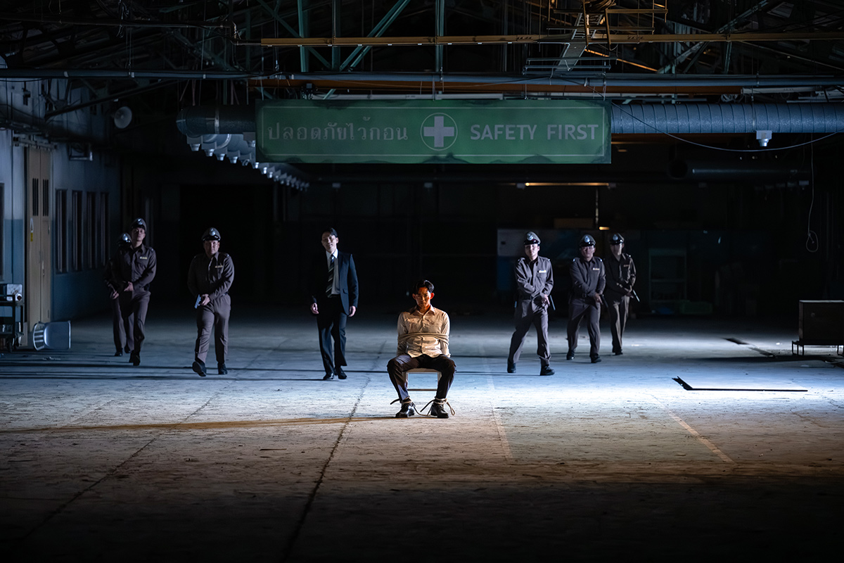 野崎の「SAFETY FIRST」の看板がある倉庫のような建物で、椅子に座った男性を制服姿の人たちが囲む場面(第14話)