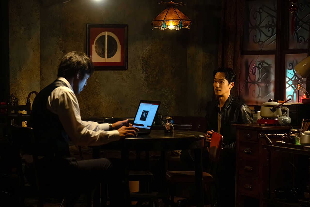 野崎の薄暗い部屋でノートパソコンに向かう男性と革のジャケットの男性が向き合う場面(第16話)