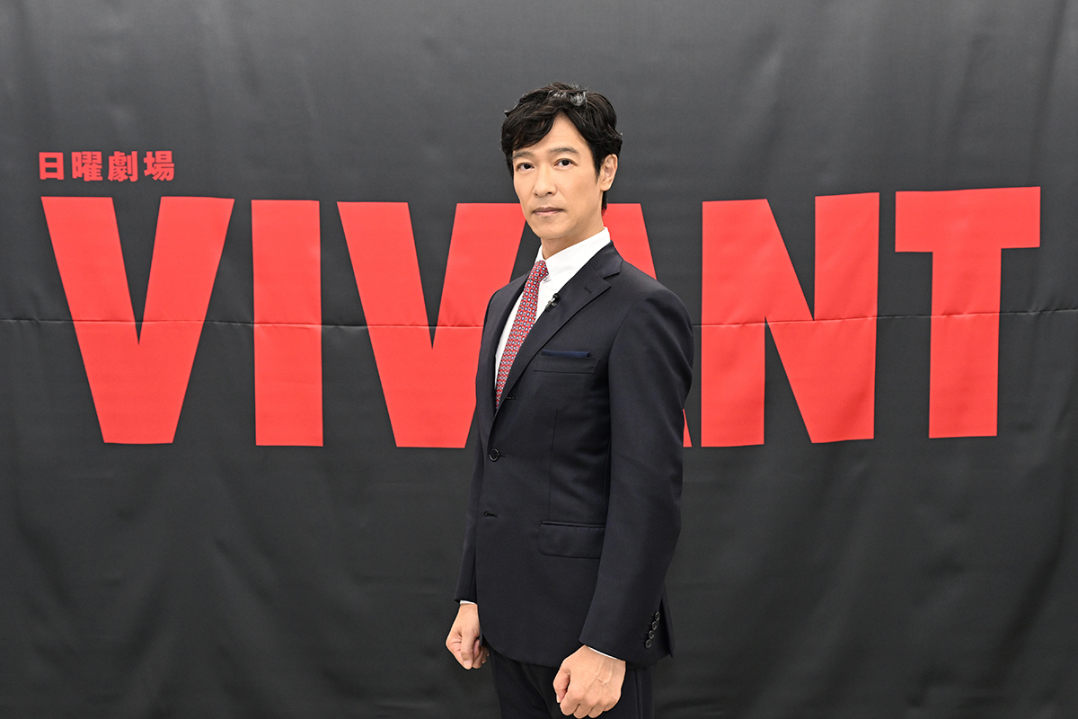 VIVANTの黒い幕に赤い大きな文字で「日曜劇場」「VIVANT」と書かれ、その前にスーツ姿の男性が立つ写真