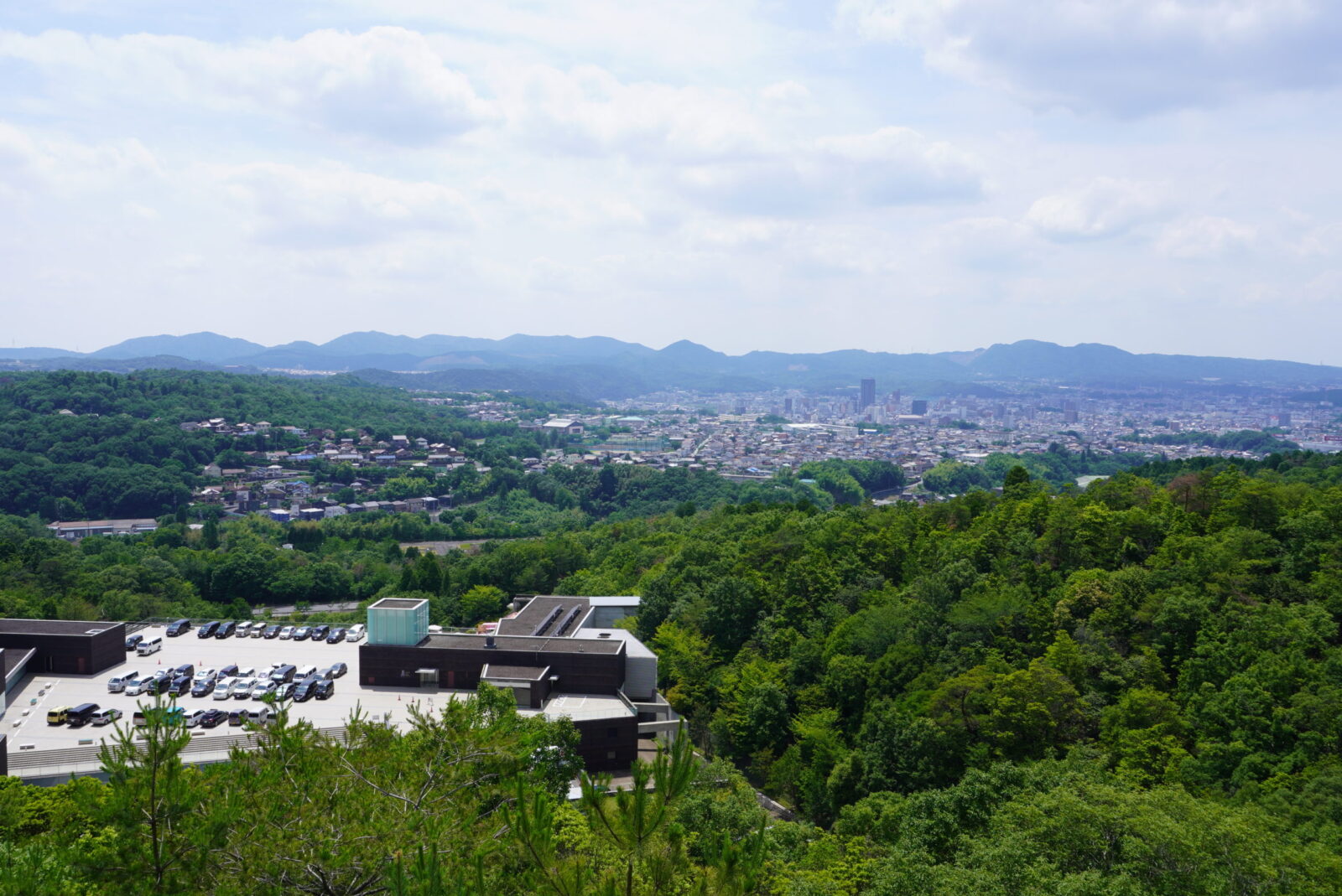 セラミックパークMINOの緑の山の中の建物と屋上の駐車場、奥に広がる街並みと山並み