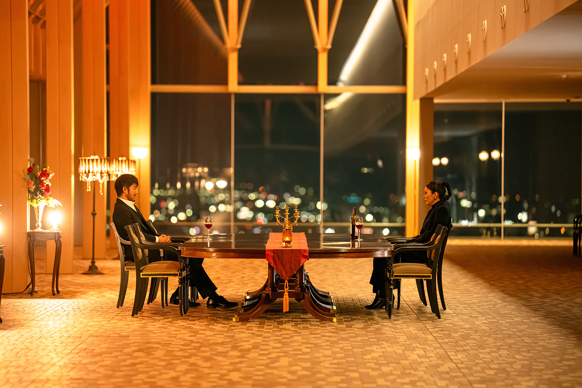 シンシーの第14話の場面写真。夜景が見える大きな窓のある広い部屋で、男女2人が長いテーブルの両端に向かい合って座る
