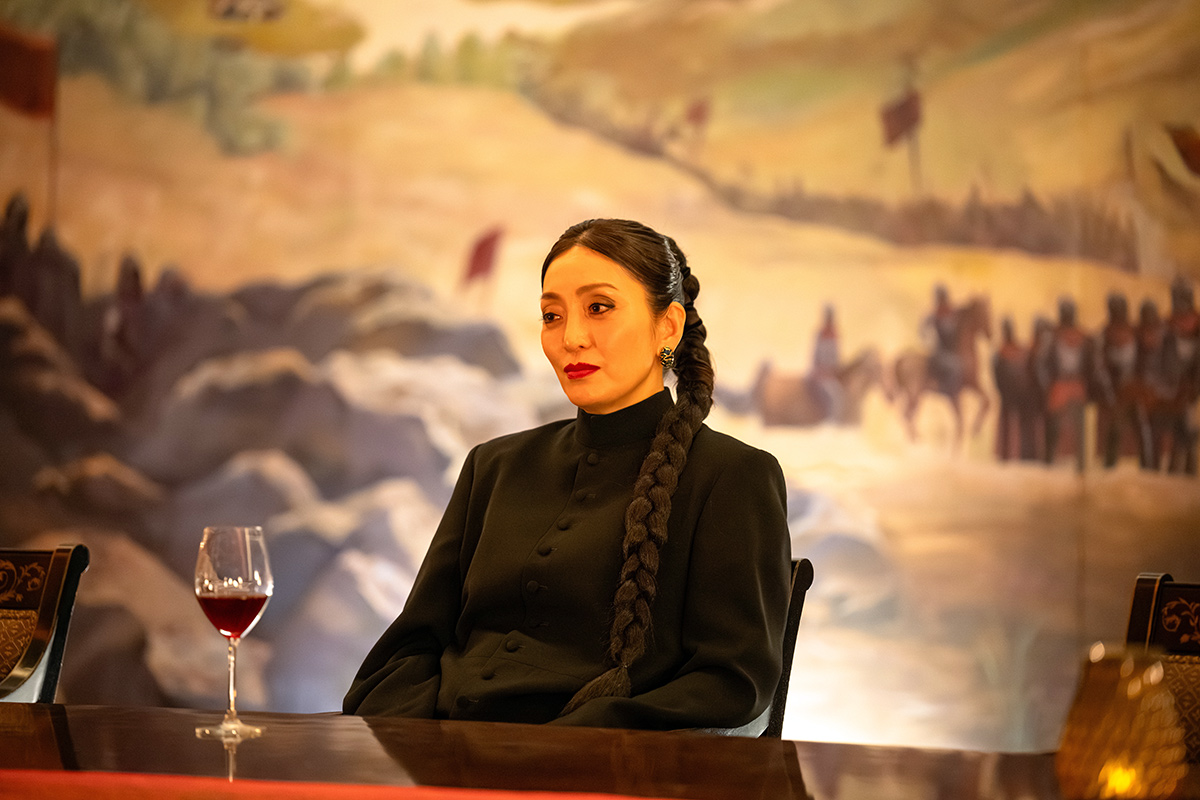 シンシーの第14話の場面写真。三つ編みの髪で黒い服の女性が、赤ワインのグラスが置かれたテーブルにつき、背後に馬に乗った人々の列を描いた大きな絵