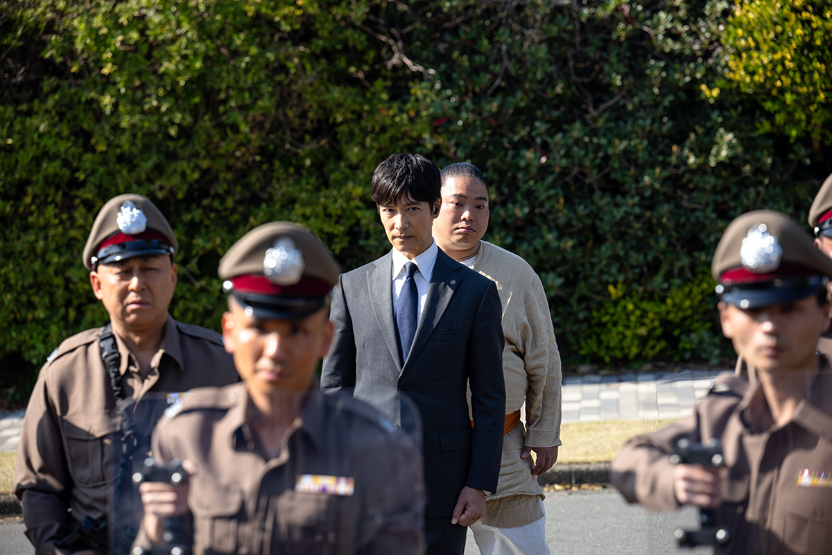 VIVANTの緑の生け垣の前の道で、制服の警官たちの後ろに立つスーツの男性とベージュの衣の大柄な人