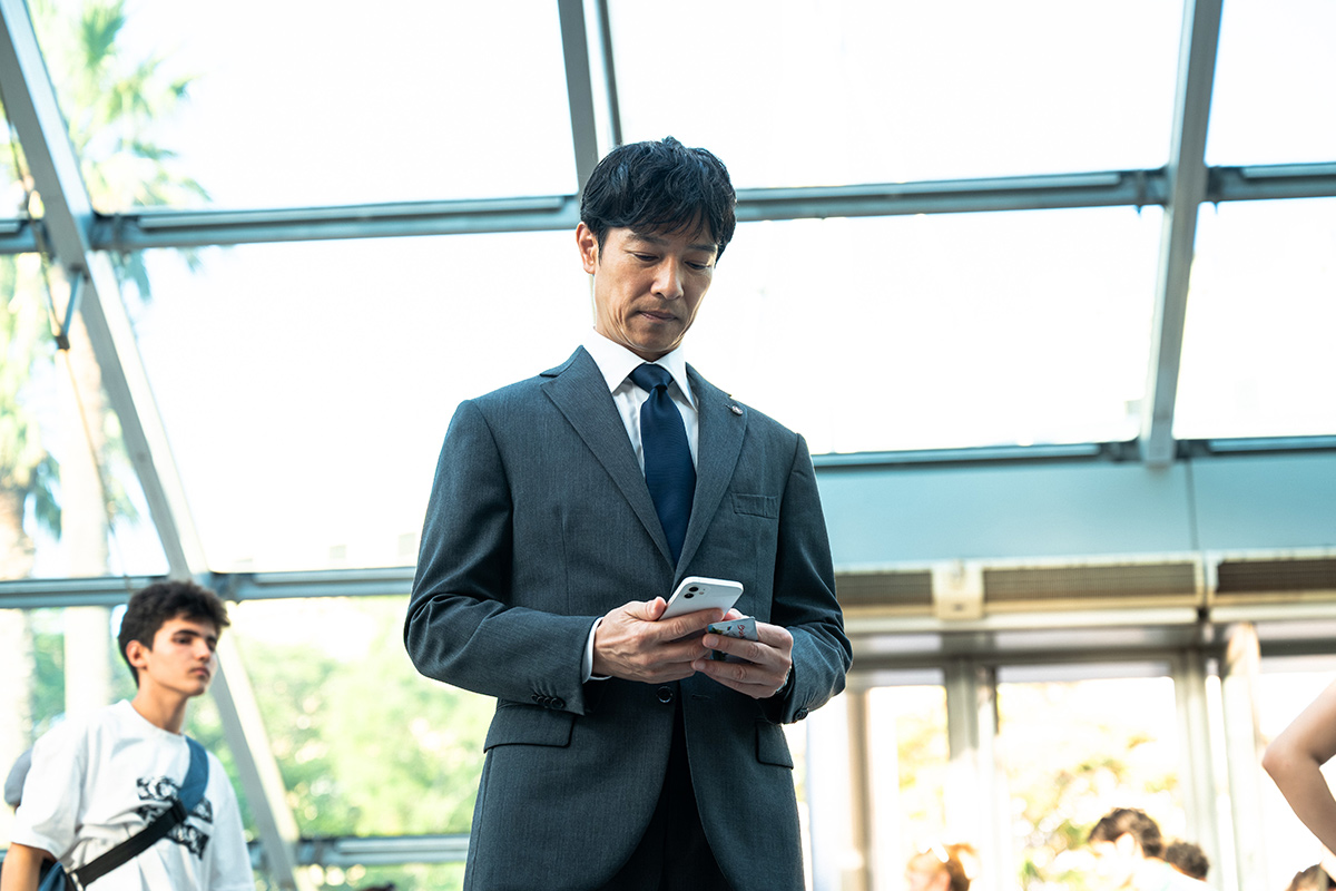 VIVANT第17話のガラス張りの天井の下で、スーツ姿の男性がスマートフォンを見る場面